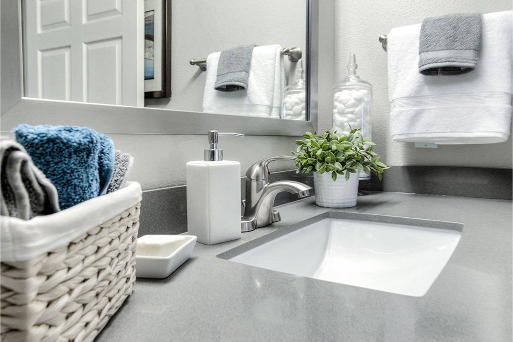 A white sink with a basket of towels and a potted plant on a grey countertop.