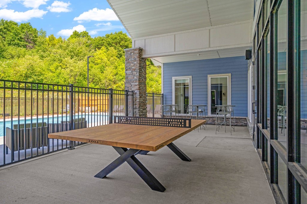 A wooden table is on a patio with a pool and trees in the background.