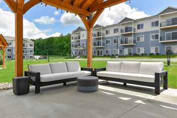 A wooden pergola with two white couches and a black ottoman in front of apartment buildings.