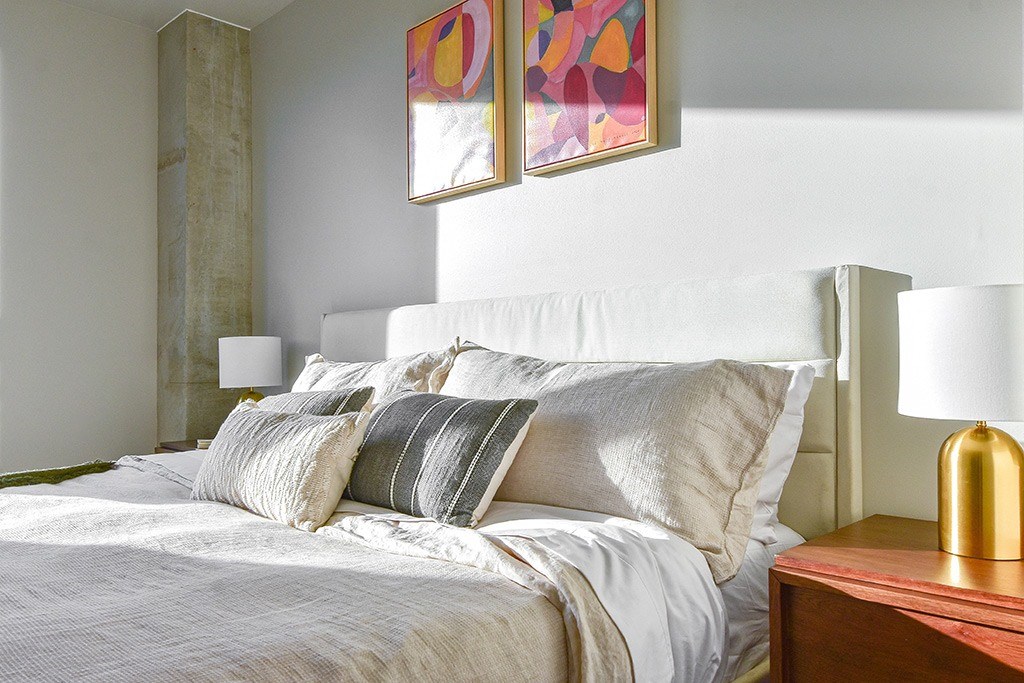 A bed with white pillows and a white comforter is in the foreground of a room with a painting on the wall.