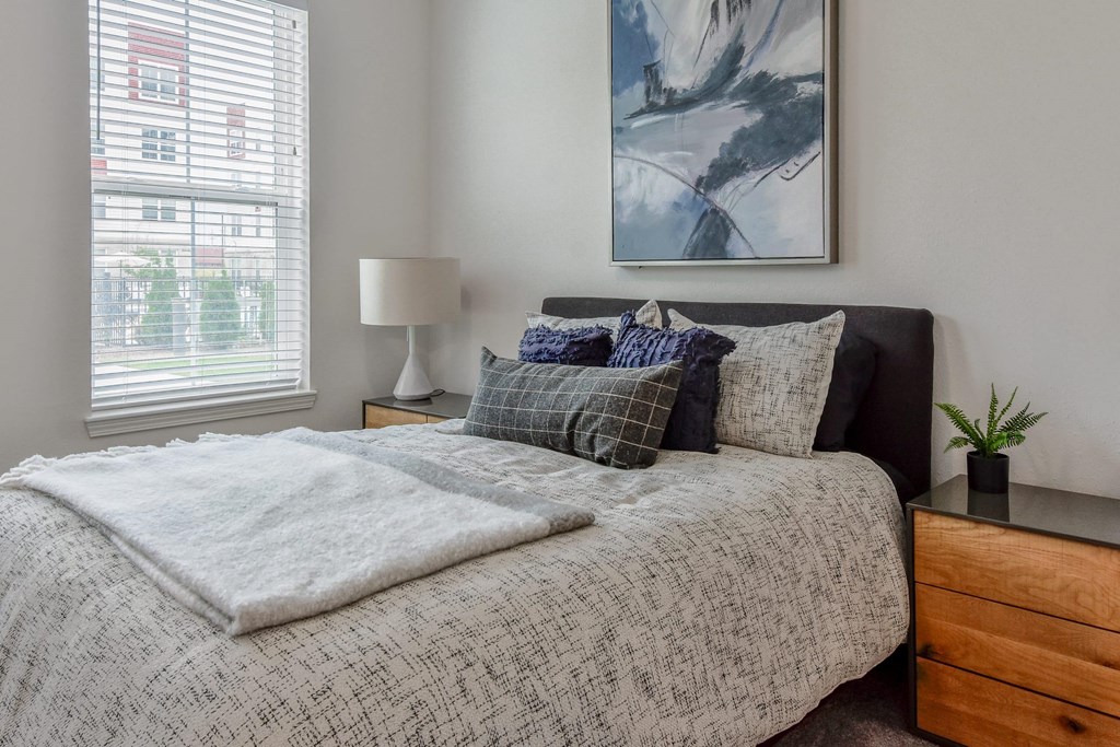 A bed with a white blanket and pillows is in front of a window with blinds.