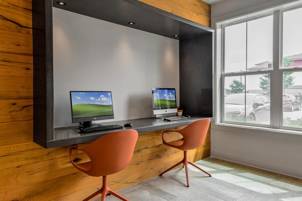 Two computers are on a desk with chairs.