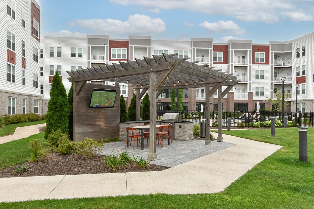 A wooden pavilion with a table and chairs is in the middle of a grassy area in front of apartment buildings.