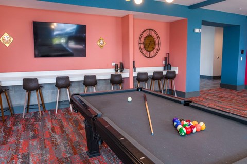 A pool table with a cue stick and balls in the foreground and a TV on the wall in the background.