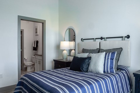 A bed with blue and white stripes is in the foreground of a room with a mirror and a lamp.