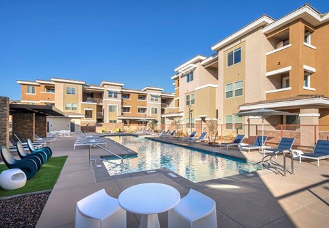 A pool area with lounge chairs and tables in front of apartment buildings.
