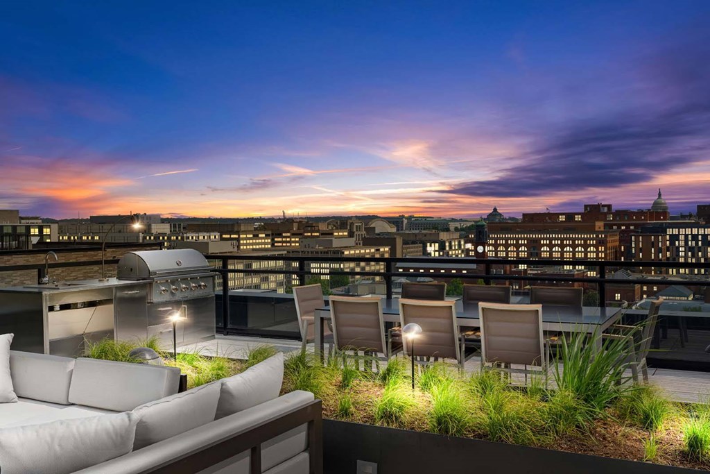A rooftop patio with a couch, chairs, and a view of the city at dusk.