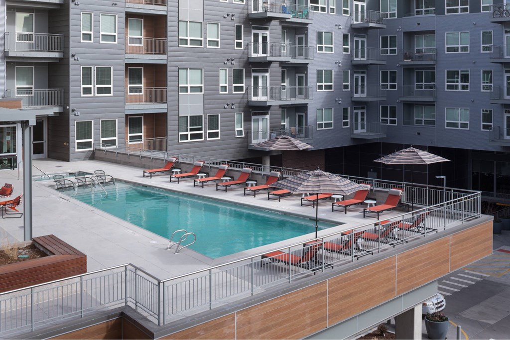 A large pool with red chairs and umbrellas is surrounded by apartment buildings.