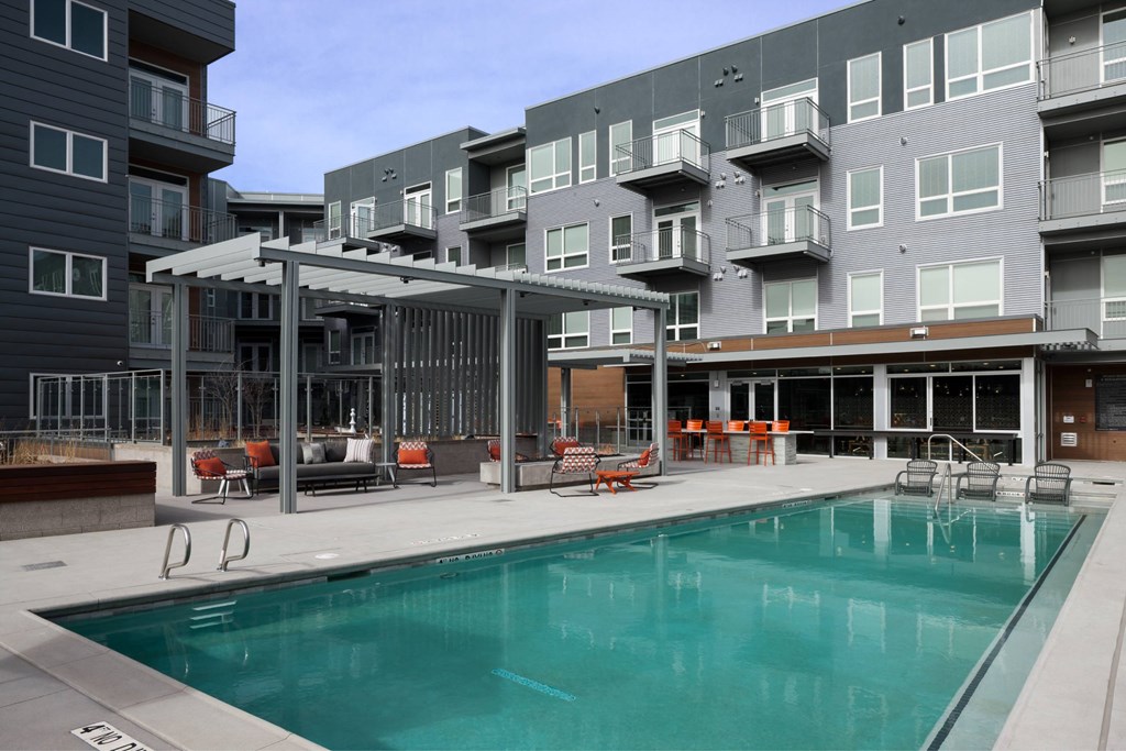 A large swimming pool is surrounded by a patio with chairs and a pergola.