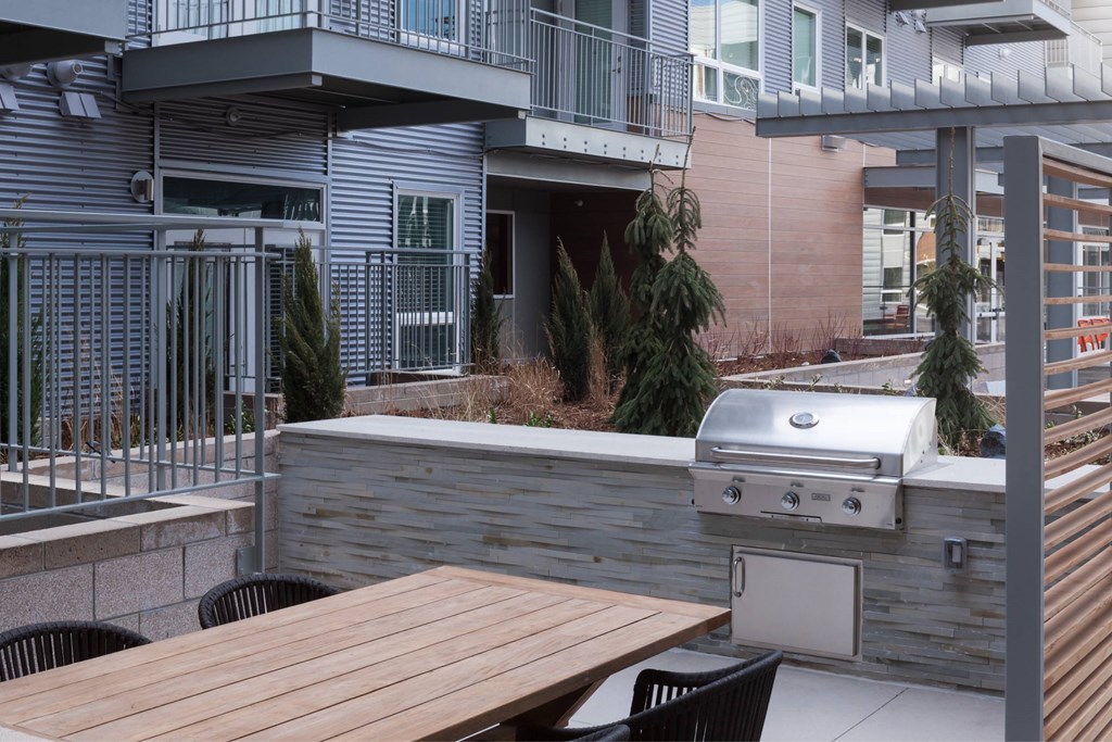 A wooden table is in front of a grill on a patio.
