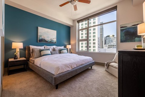 A bedroom with a bed, a ceiling fan, and a window overlooking a cityscape.