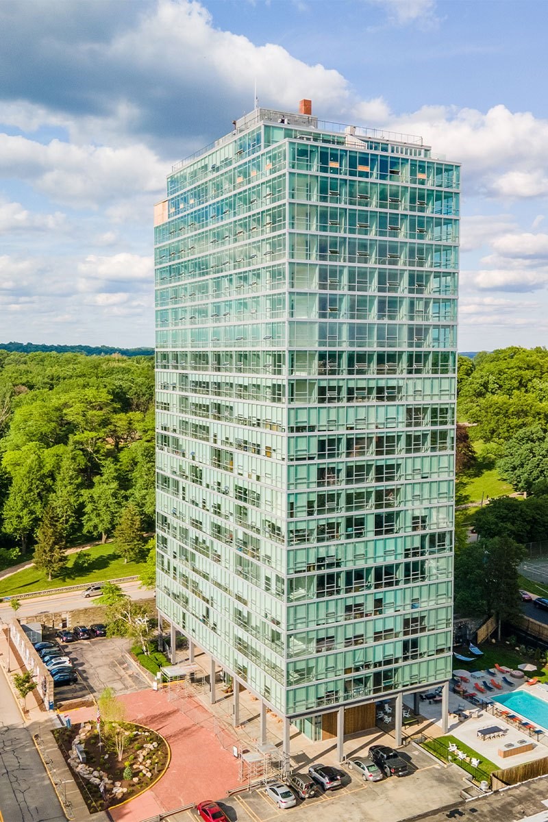 A tall glass building with a red roof is surrounded by trees and has a parking lot in front.