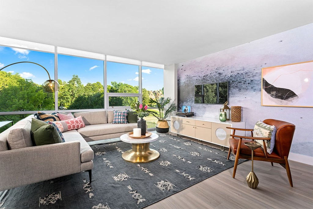 A living room with a grey couch, a round gold coffee table, and a large window with a view of trees.
