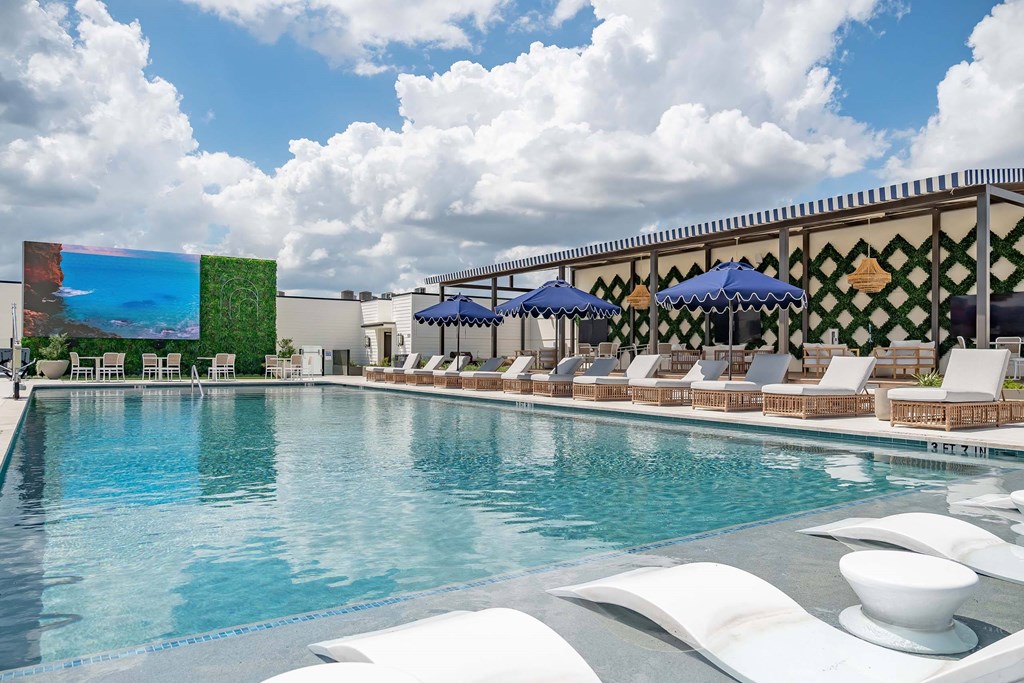 A pool with white chairs and umbrellas is surrounded by a building with a green wall.