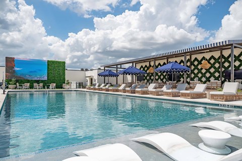 A pool with white chairs and umbrellas is surrounded by a building with a green wall.