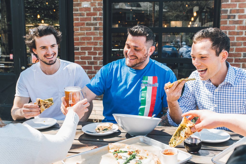 Four people eating pizza and drinking beer at an outdoor table.