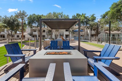 A fire pit is surrounded by blue chairs and benches in a sunny outdoor setting.