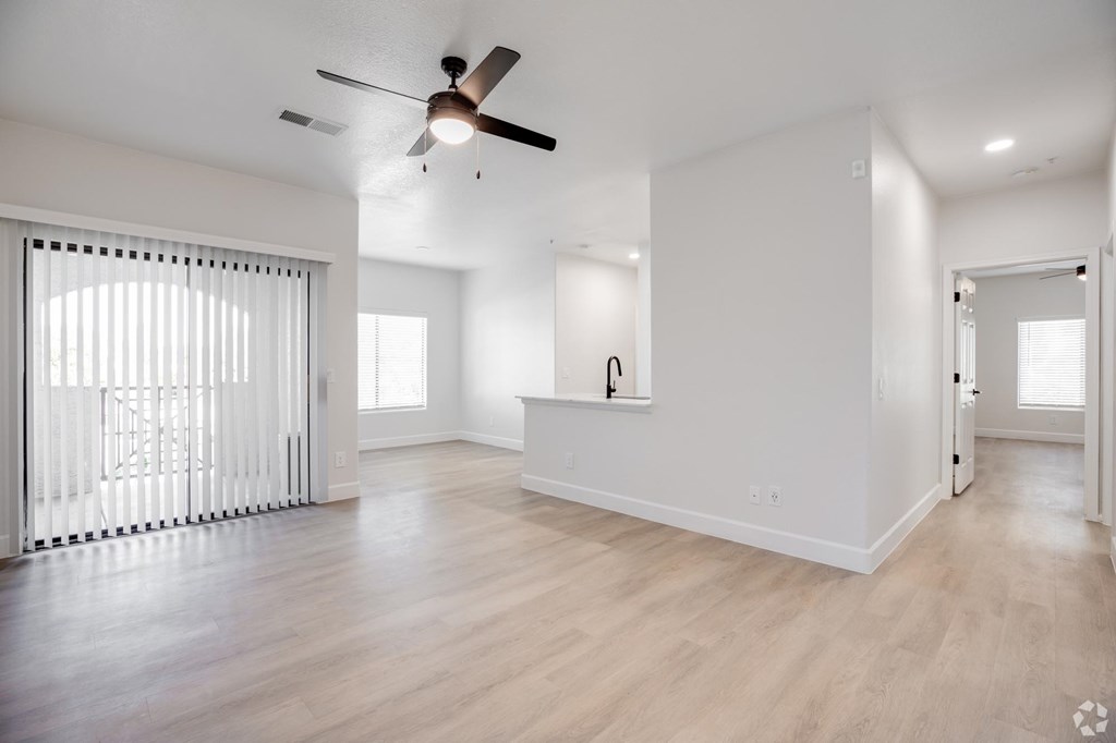A spacious room with a ceiling fan and light fixture, hardwood flooring, and a window with blinds.