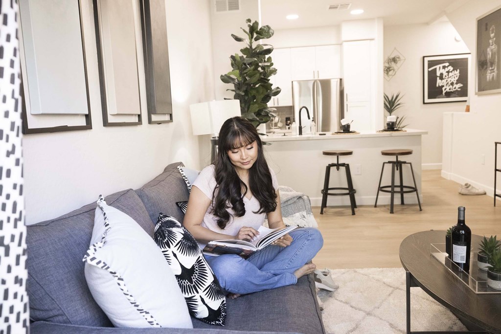 A woman is sitting on a grey couch reading a book.