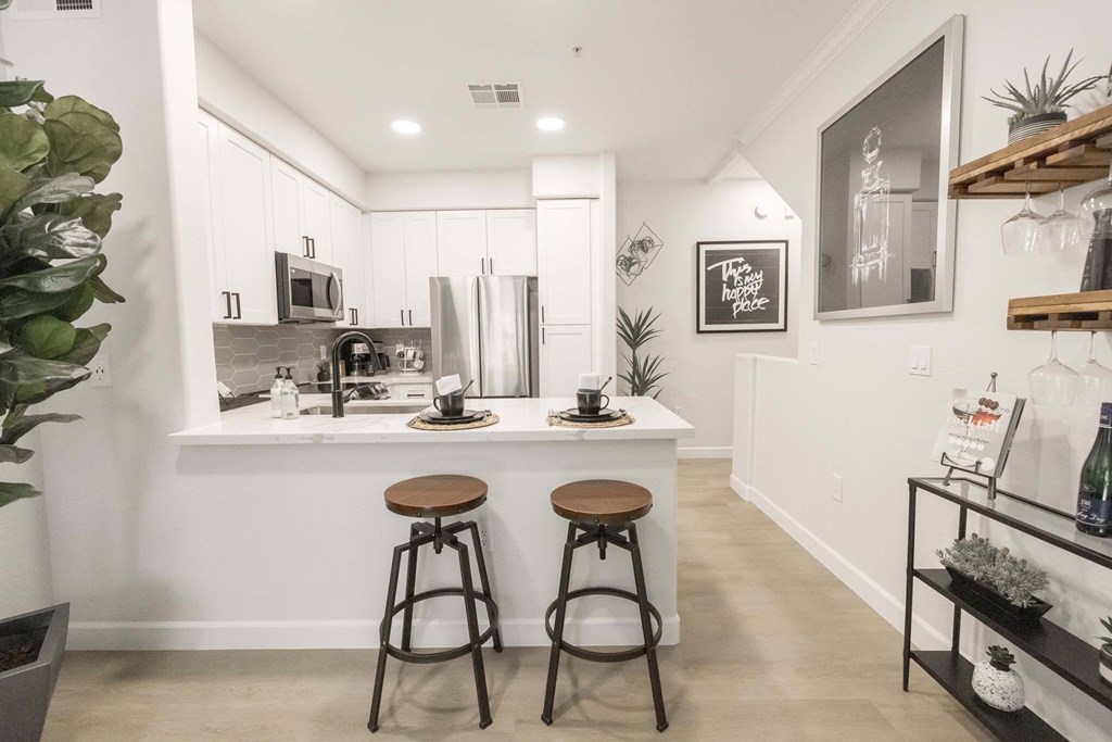 A kitchen with white cabinets and a white countertop with two stools in front of it.