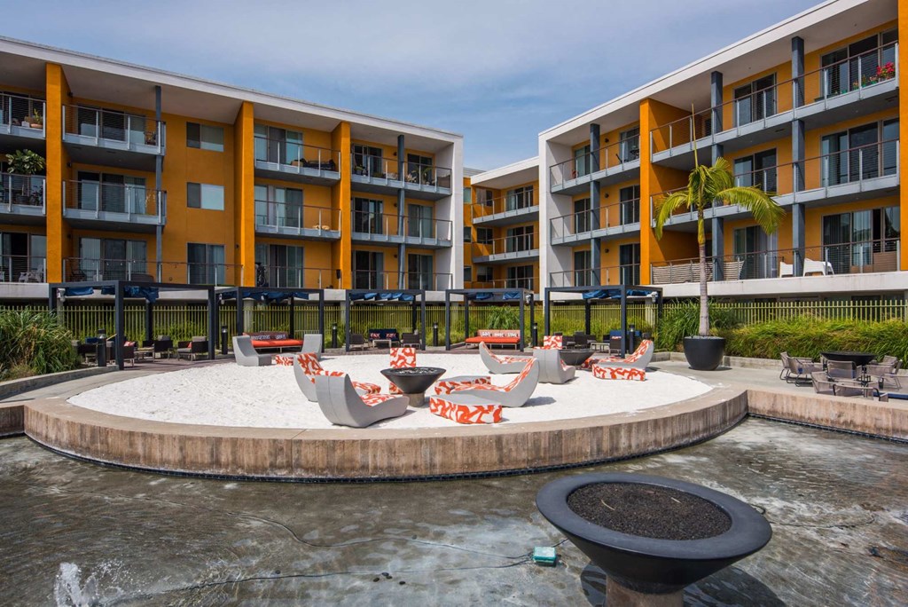 A round sand pit is surrounded by chairs and tables in front of apartment buildings.