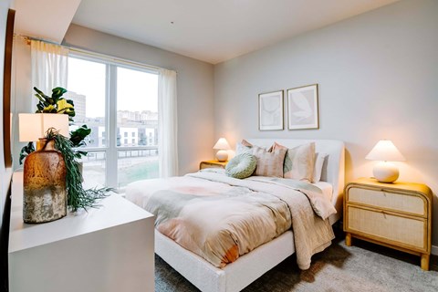 A bedroom with a large bed, a nightstand, and a window with a view of the city.