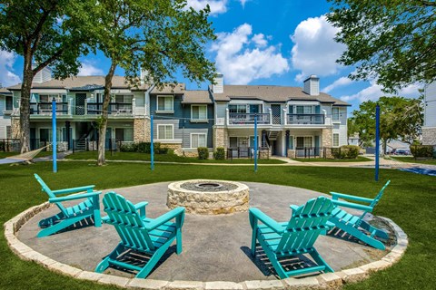 A fire pit is surrounded by four blue Adirondack chairs in a grassy area.