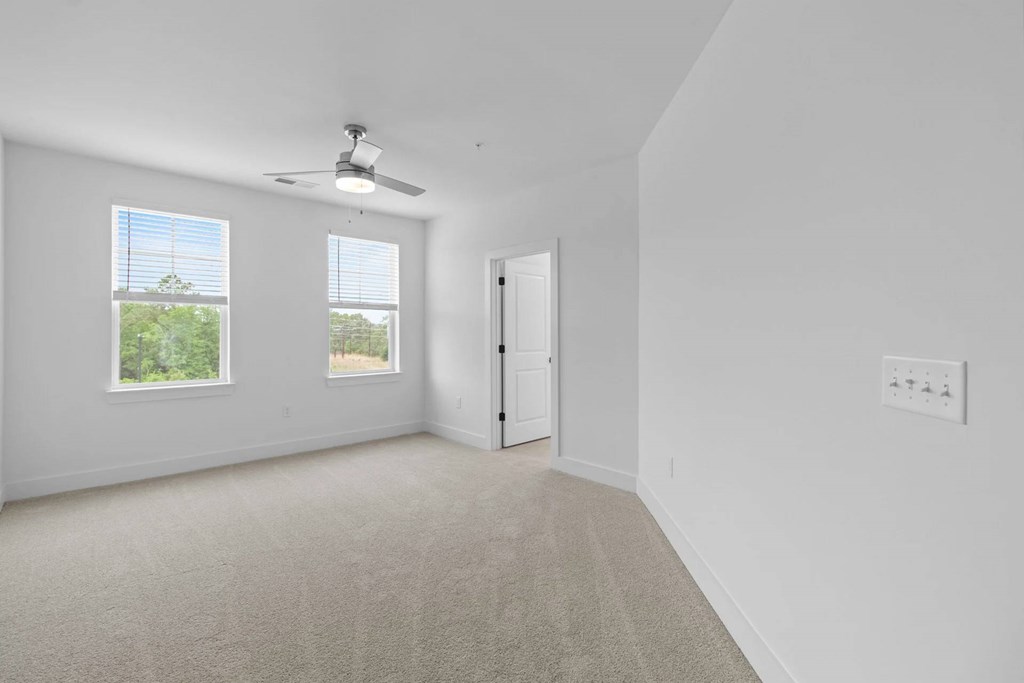 A room with a carpet floor, a ceiling fan, and a door leading to another room.