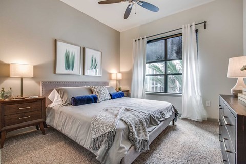 A bedroom with a large bed, a ceiling fan, and a window with white curtains.