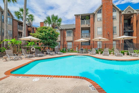 A swimming pool surrounded by lounge chairs and umbrellas in front of a brick building.