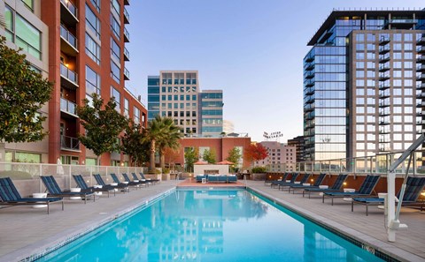 A large swimming pool is surrounded by lounge chairs in front of apartment buildings.
