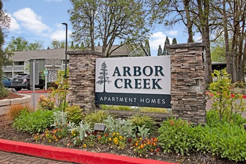 A sign for Arbor Creek Apartment Homes is surrounded by flowers and plants.