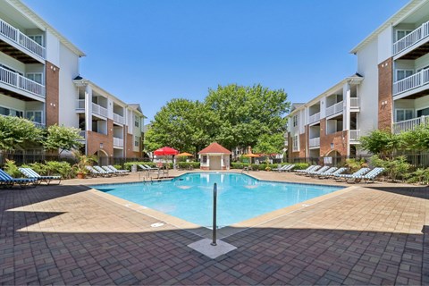 A swimming pool surrounded by a brick patio and lounge chairs with a red umbrella and apartment buildings in the background.