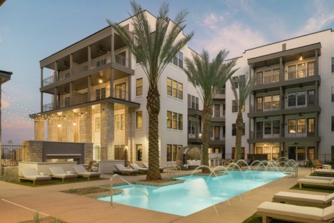 A swimming pool is surrounded by lounge chairs and palm trees in front of apartment buildings.