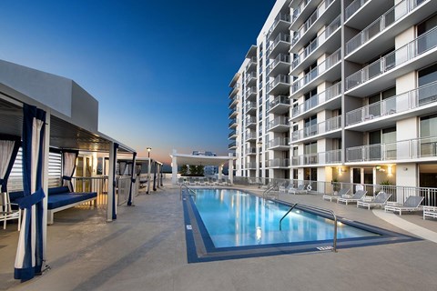 A swimming pool is surrounded by lounge chairs and apartment buildings.