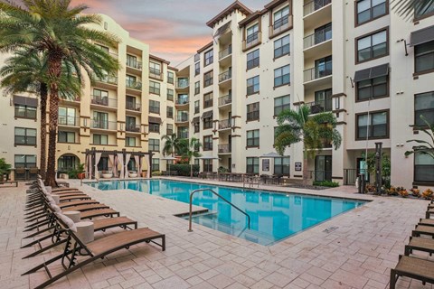 A large swimming pool surrounded by sun loungers and palm trees in front of apartment buildings.