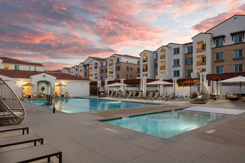 A large swimming pool surrounded by lounge chairs and umbrellas in front of apartment buildings.