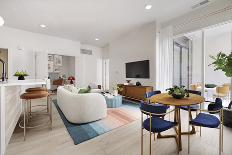 A modern living room with a white sofa, a round table with blue chairs, and a kitchen area in the background.