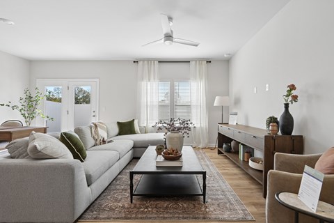 A living room with a grey sofa, a coffee table, a ceiling fan, and a window with white curtains.