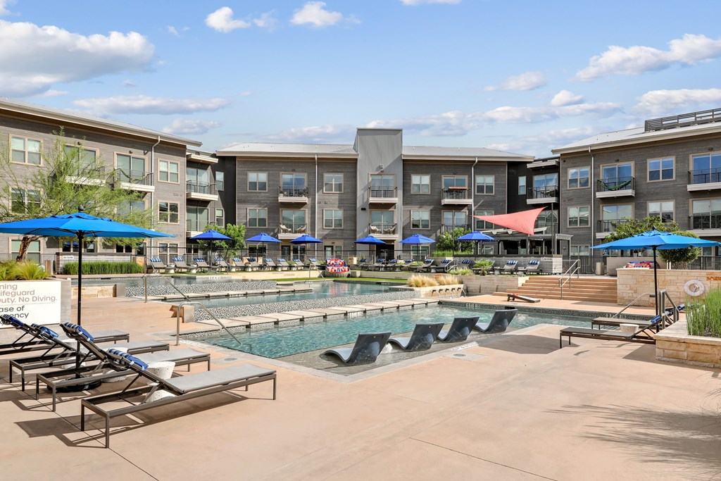 A swimming pool area with sun loungers and umbrellas in front of apartment buildings.