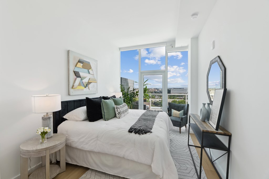 A bedroom with a large bed, a side table with a lamp, and a view of the city through the window.