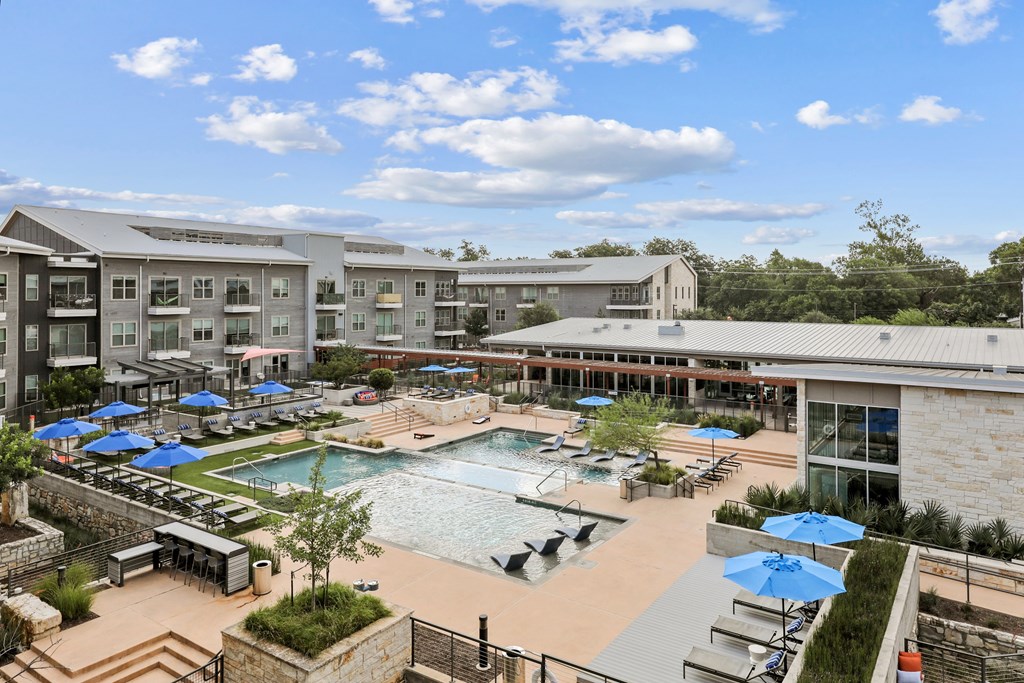 A large outdoor swimming pool with loungers and umbrellas is surrounded by apartment buildings.