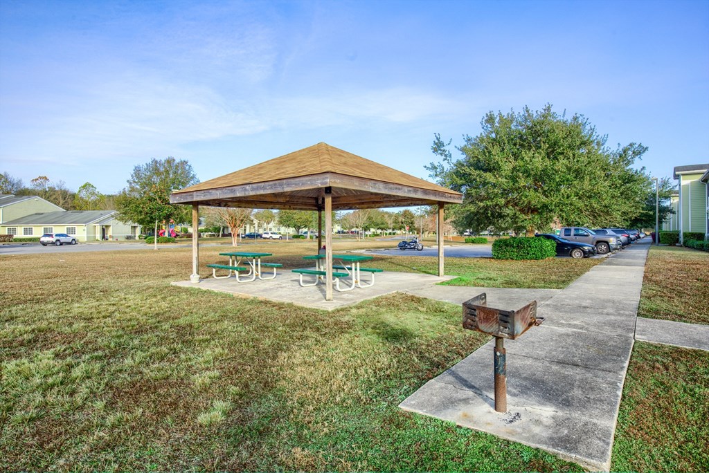 A pavilion with a wooden roof and a picnic table is surrounded by a grassy area.