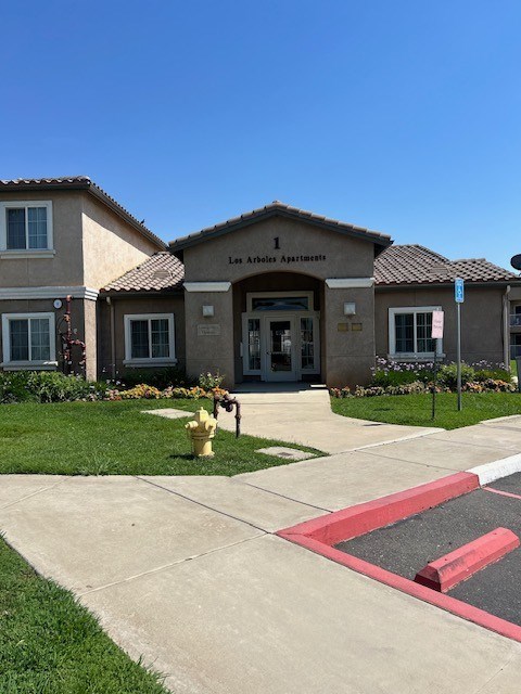 The image shows the front of the Los Arboles Apartments with a clear blue sky in the background.