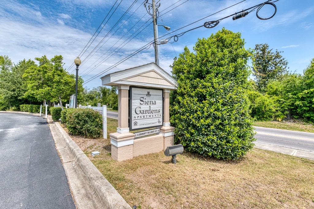 A sign for Siena Gardens is situated on a grassy area next to a road.