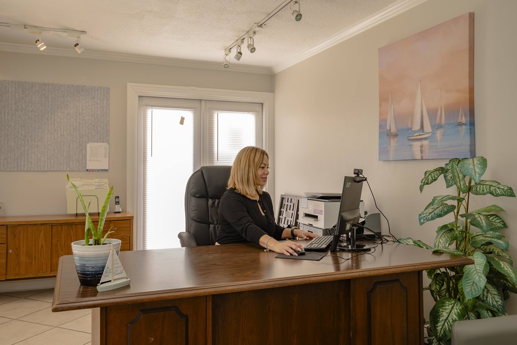 A woman is sitting at a desk in an office with a computer and a plant.