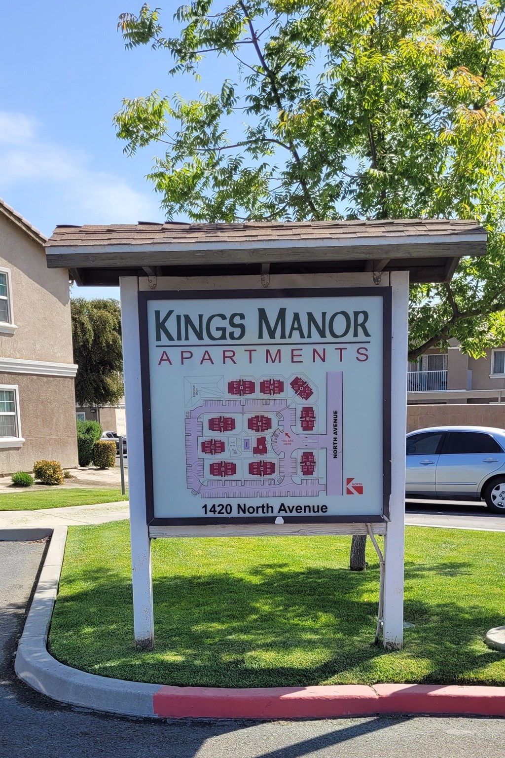 A Kings Manor Apartments sign is in front of a tree and a building.