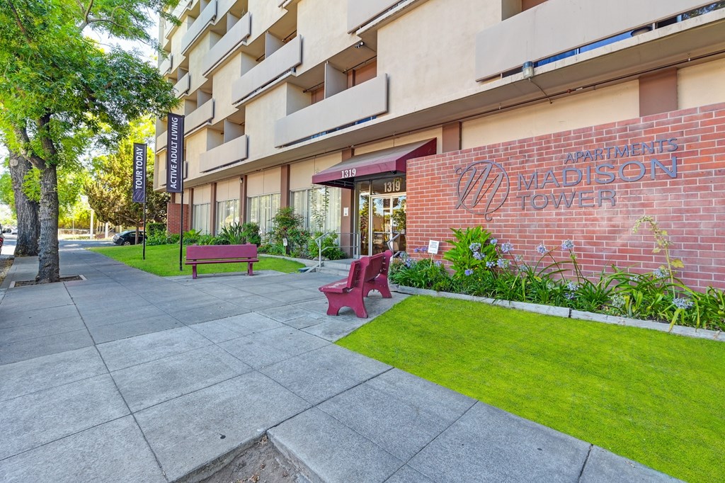 A red bench sits on a sidewalk in front of a building named Madison Tower.