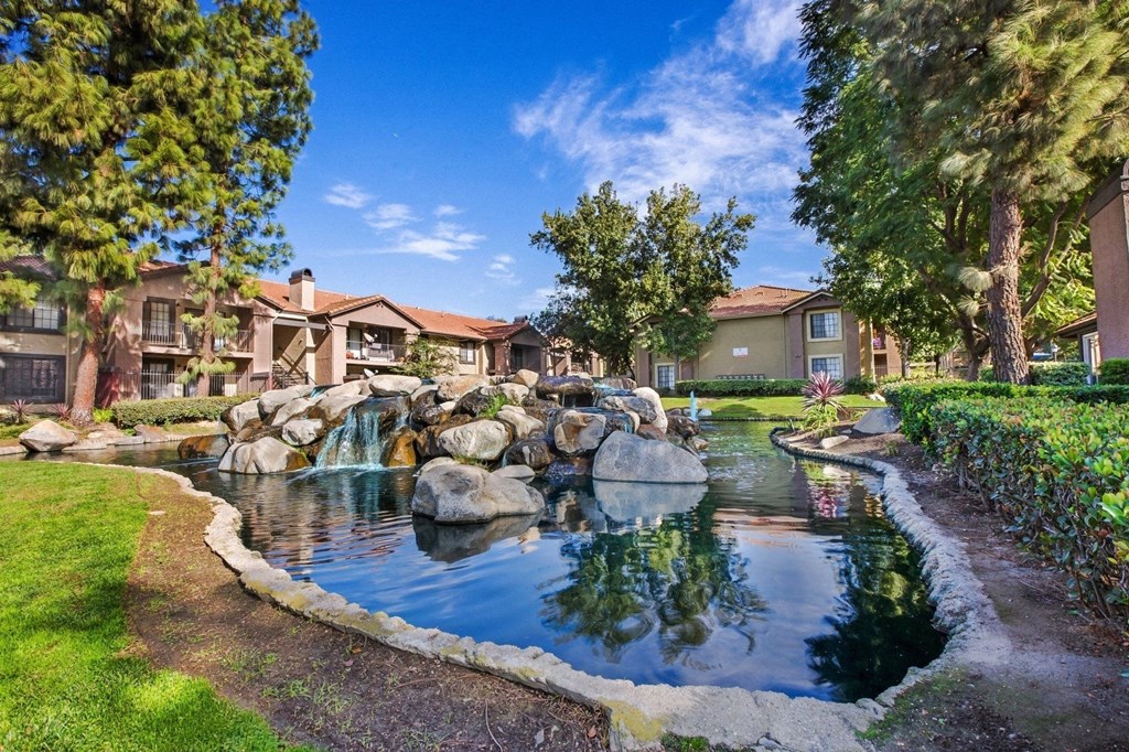 a fountain in the middle of pond surrounded  a grassy area with buildings in the background  at Citrine Hills, Ontario, CA, 91761
