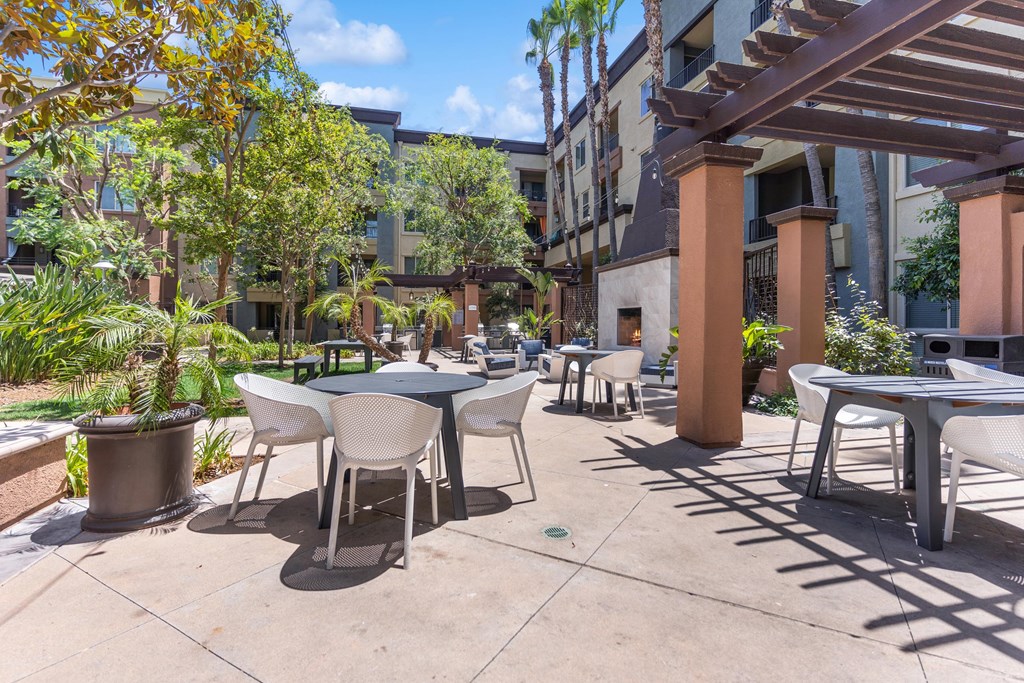 A patio with tables and chairs is surrounded by plants and trees at The Kitt at Warner Center Apartments, Woodland Hills 91303